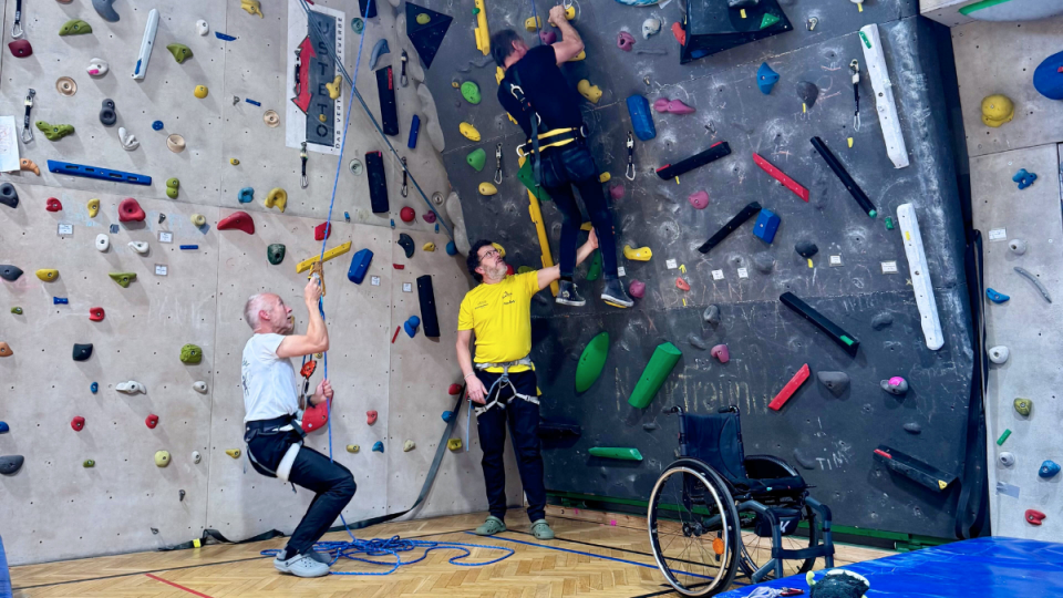 2 Klettertrainer erklären einem Mann im Rollstuhl die Sicherungstechniken beim Klettern